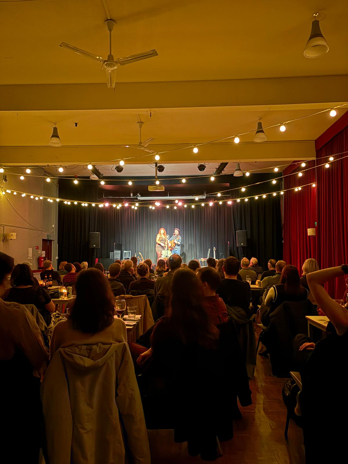 Audience seated beneath string lights watching A Different Thread at Bristol Folk House